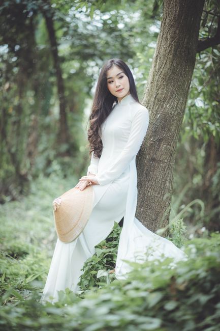 Asian woman in traditional dress leaning against a tree, holding a hat in a natural setting.