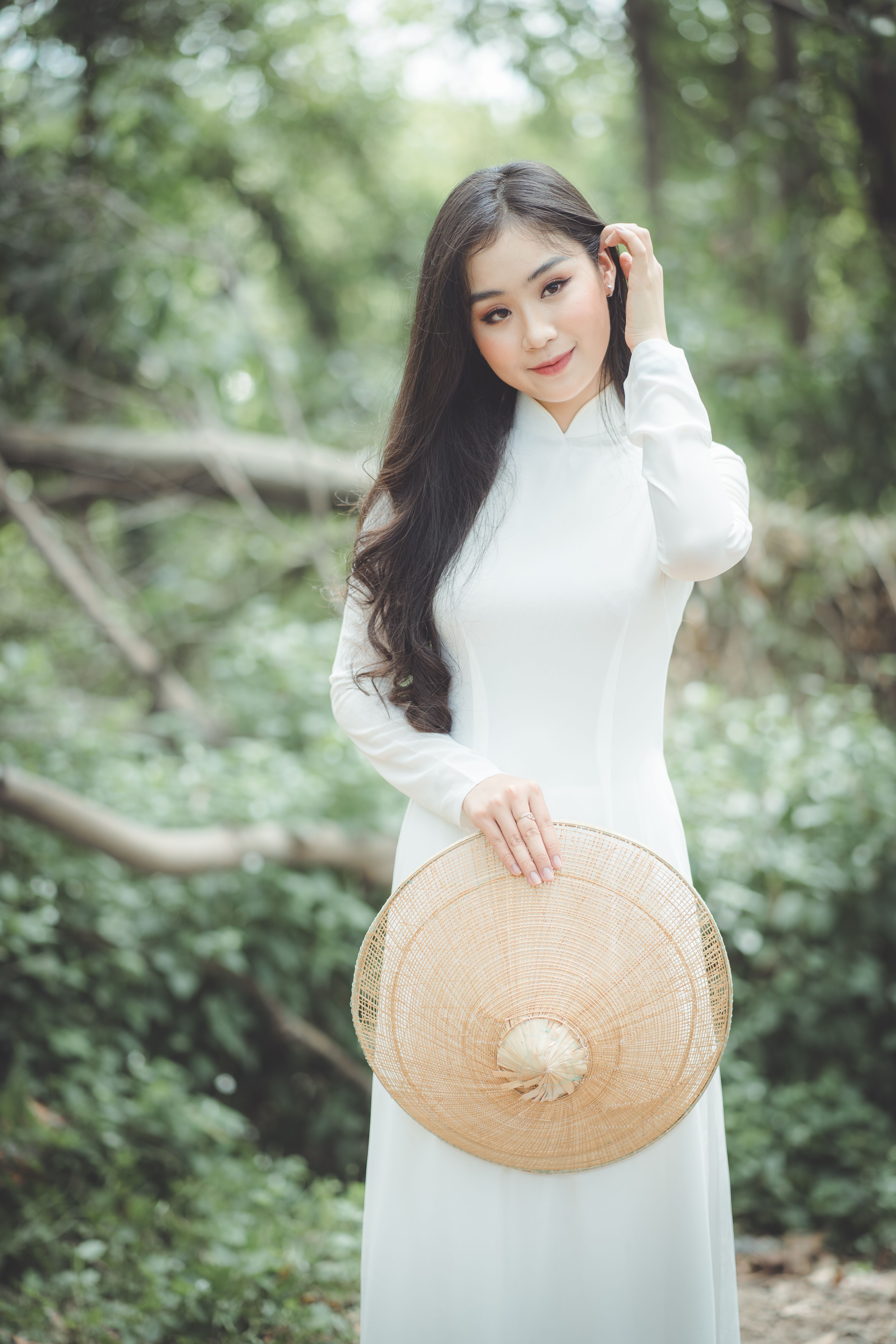 Elegant Asian woman in a white ao dai with a conical hat in a serene outdoor setting.