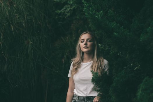 Portrait of a young woman in a white shirt standing amidst lush green foliage, outdoors.