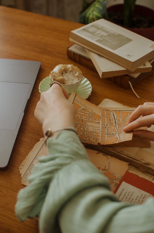 High angle of crop faceless lady in blouse at table with old maps and papers near netbook and cup with coffee on saucer near books in light room