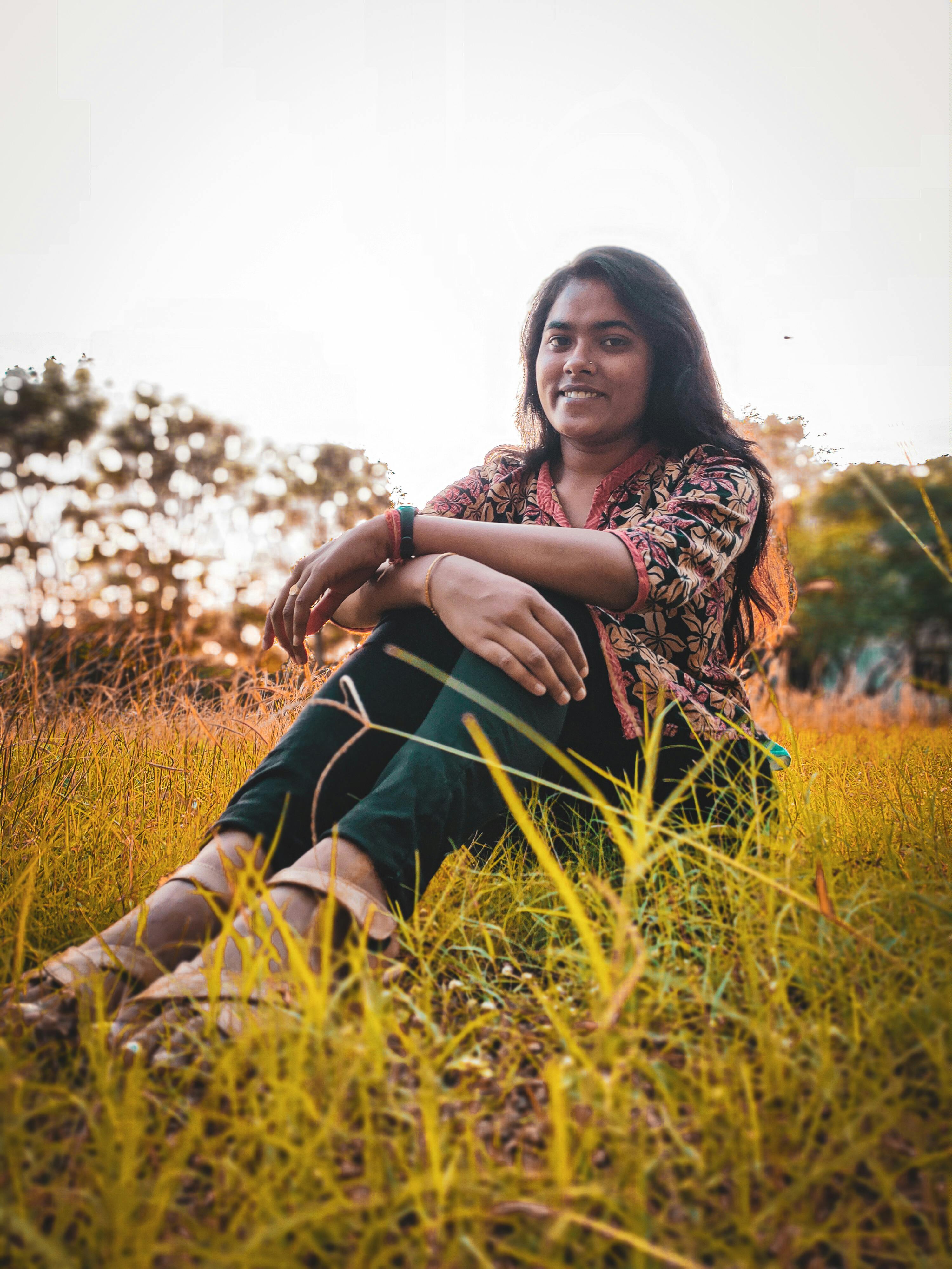 A cheerful woman sits in a grassy field, enjoying a serene sunset. Captured outdoors in India.