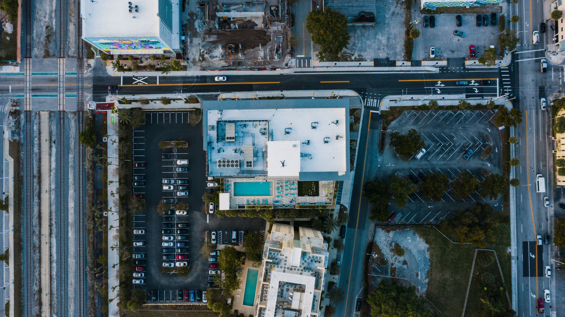 Drone view of modern town with buildings near parking lot with green trees near asphalt roads with vehicles in daylight