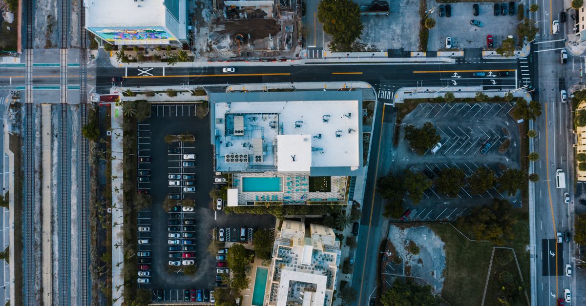Drone view of modern town with buildings near parking lot with green trees near asphalt roads with vehicles in daylight