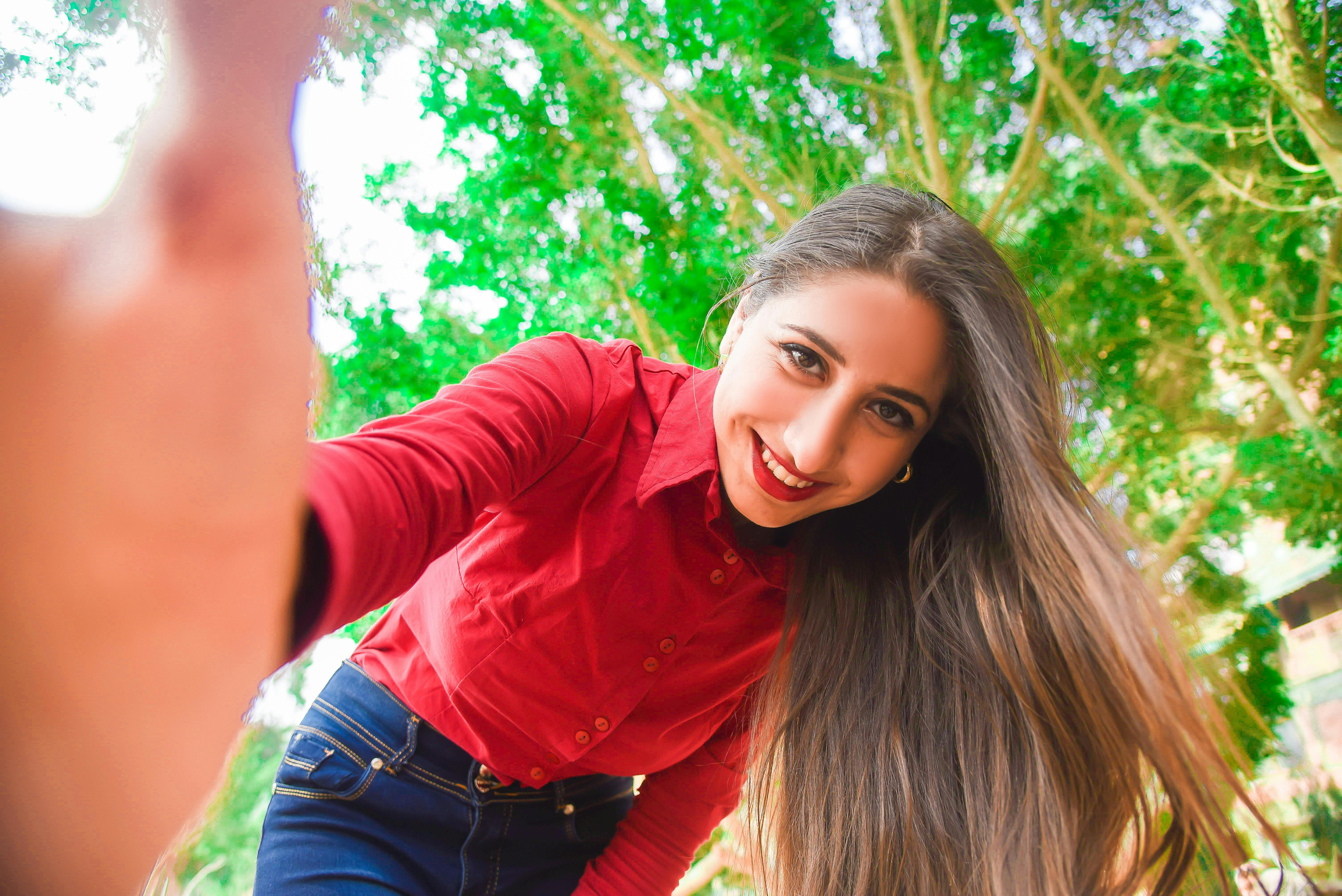 Smiling Woman in Red Shirt and Blue Jeans Taking Selfie Under Green Leaved Tree \u00b7 Free Stock Photo