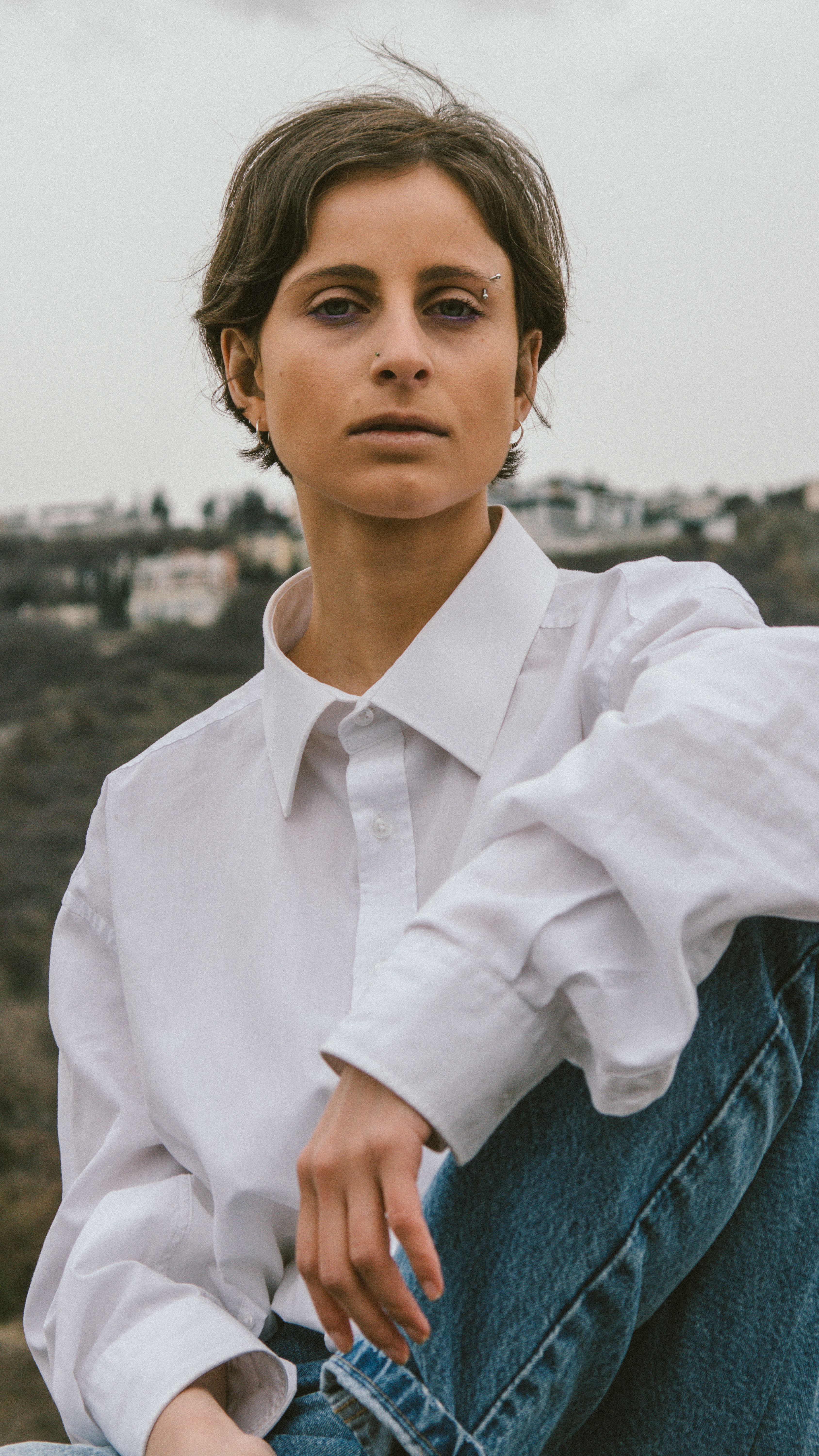 Woman with eyebrow piercing, wearing a white shirt and jeans outdoors.