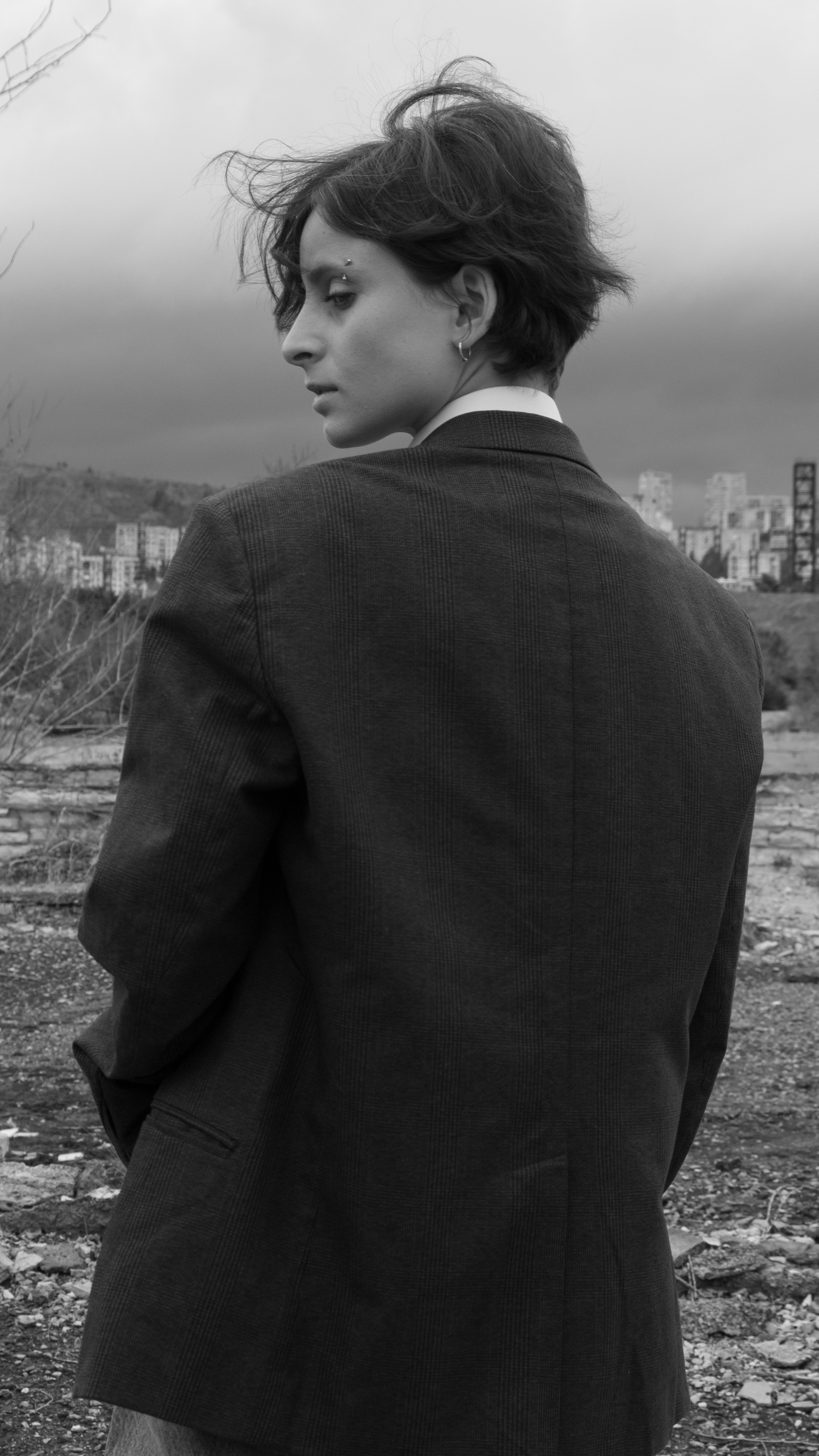 A contemplative woman in a jacket stands outdoors, facing away, in an urban landscape.