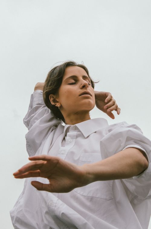 Free A woman in a white button-down shirt poses expressively against a muted sky, showcasing modern fashion. Stock Photo
