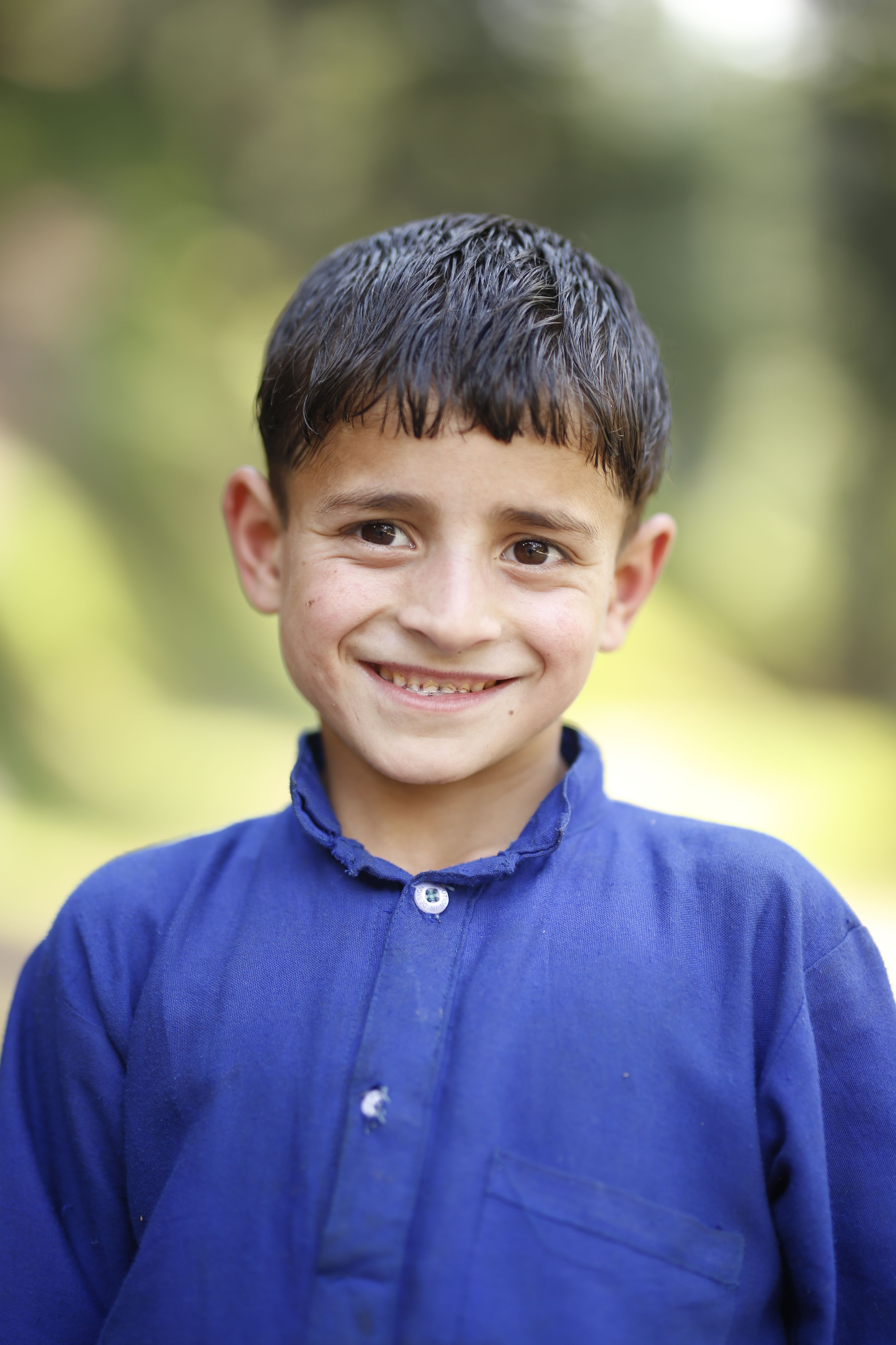 A cheerful child smiles warmly in a vibrant blue shirt, set against a lush outdoor backdrop.