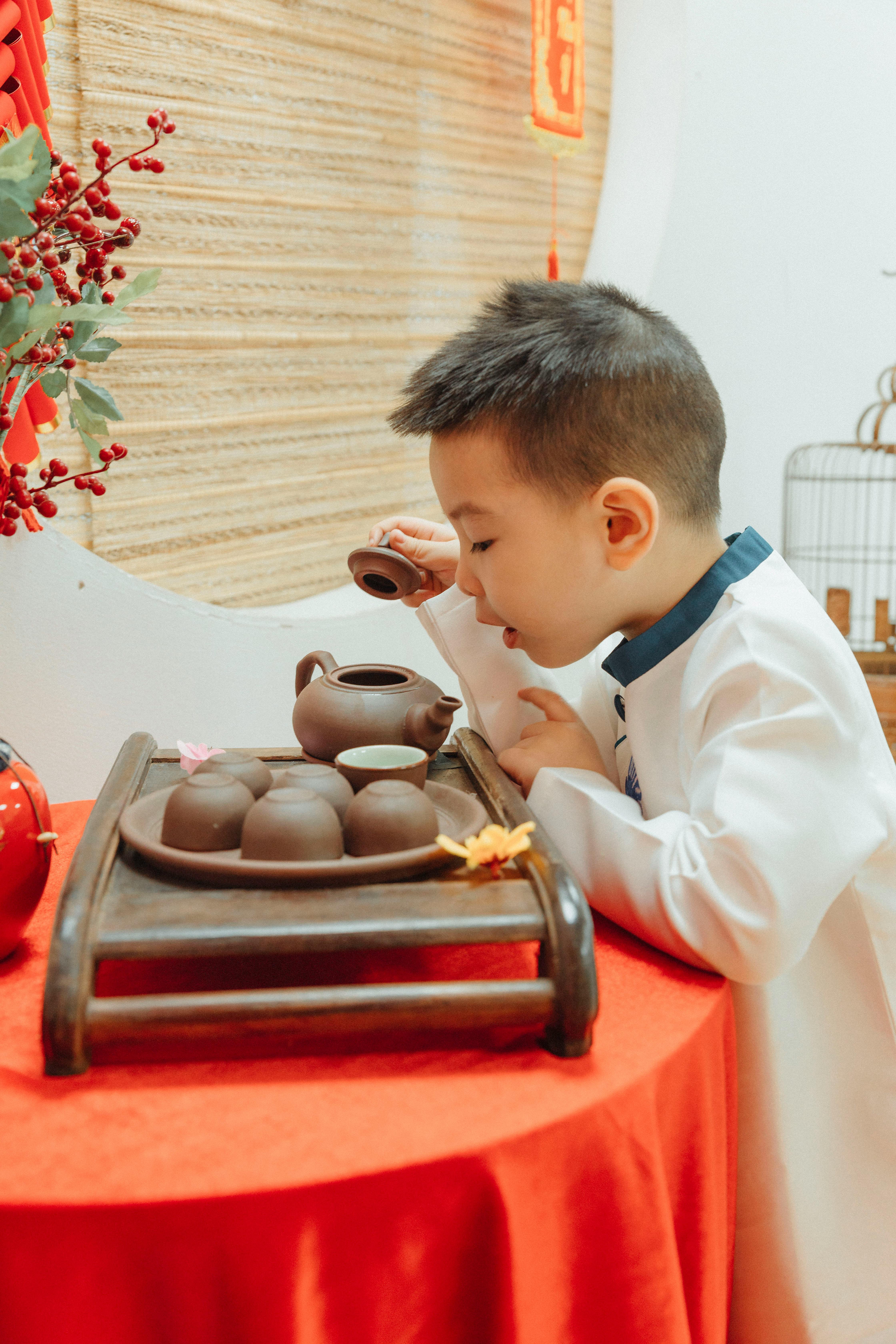 A young boy in traditional dress performing a tea ceremony with a teapot on a red table indoors.