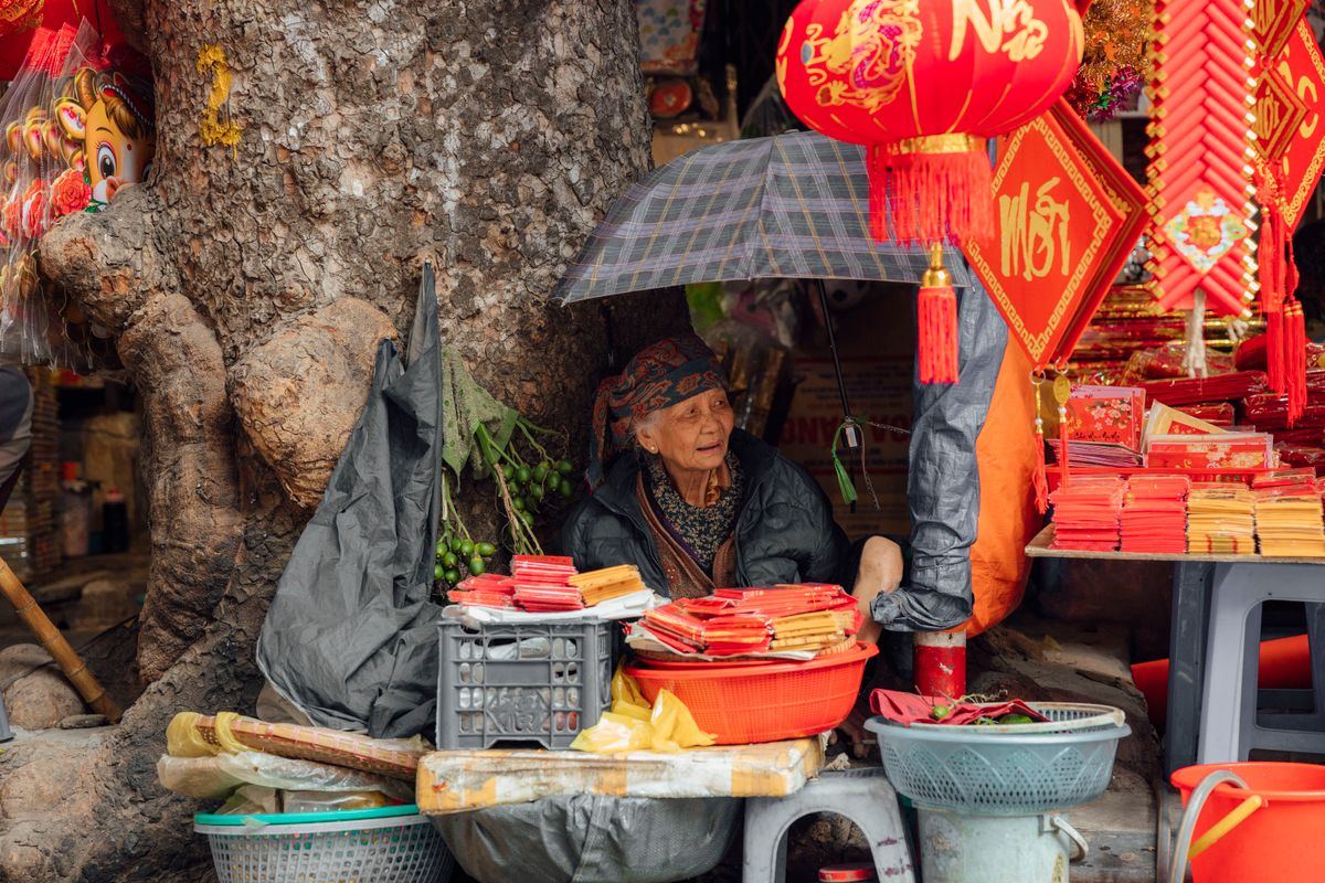 Elderly Chinese woman in traditional clothing seated at a street stall selling red decorations