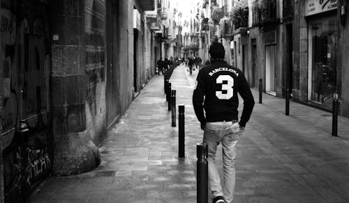 Free Grayscale Photo of Man Wearing Long-sleeved Shirt and Pants Walking on Concrete Pathway Near Building Stock Photo