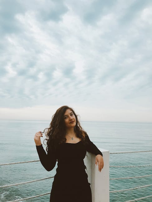 Woman in a black dress posing by the sea with cloudy sky backdrop.