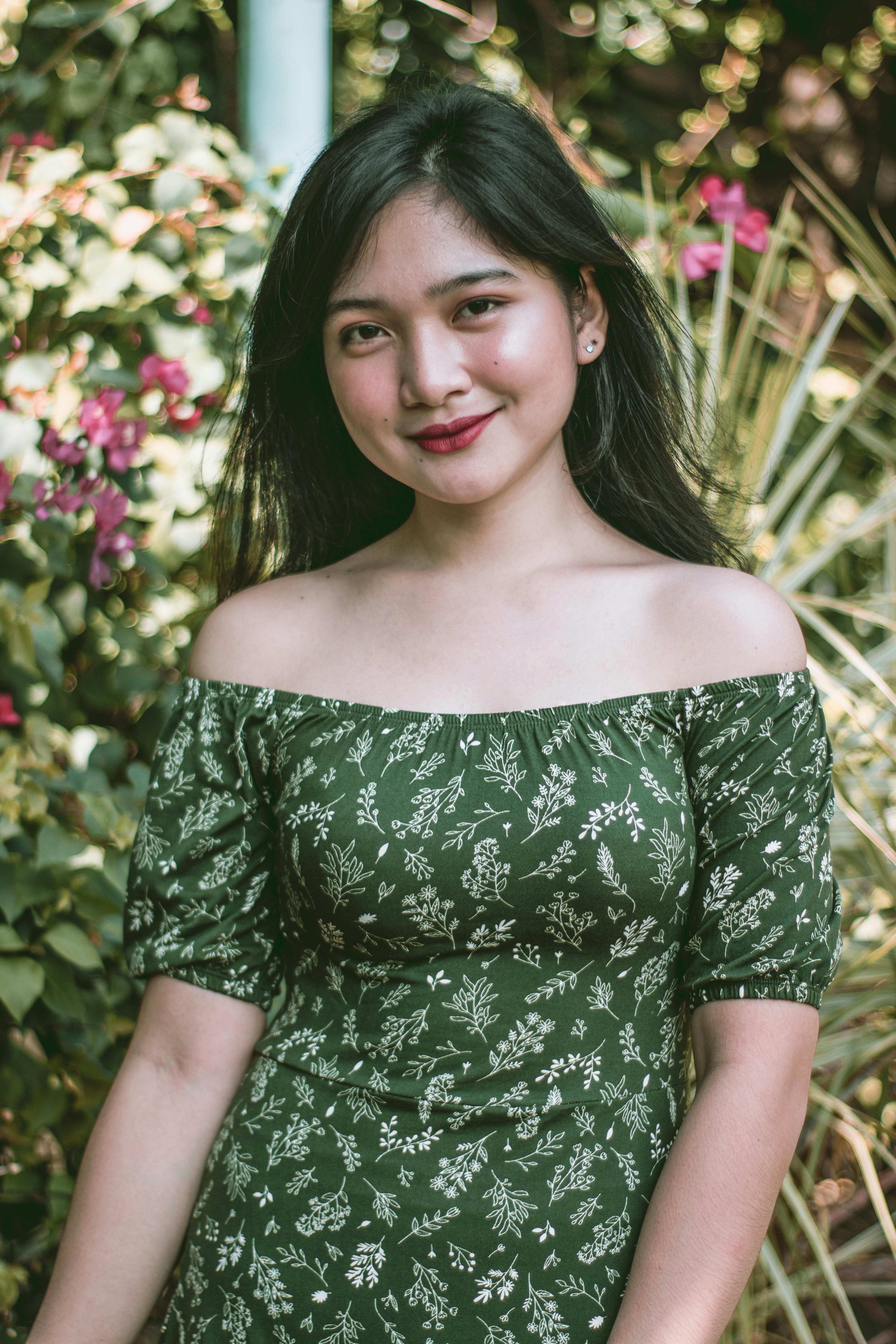 A smiling woman in a green floral dress stands outdoors with a blurred natural background.