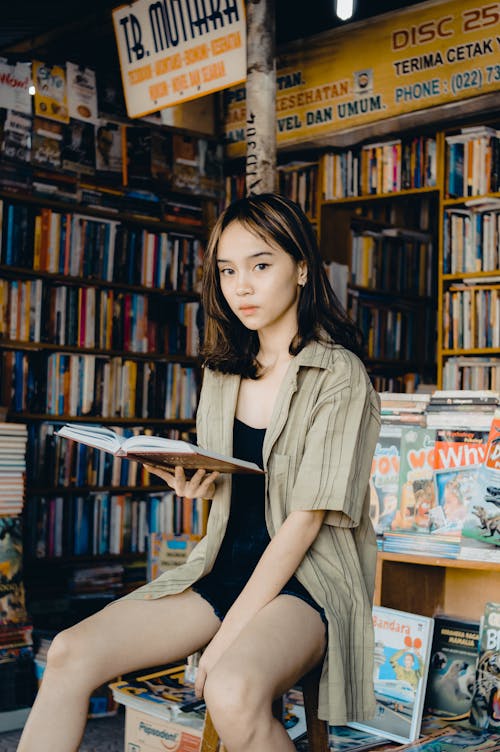 Free Pensive ethnic female in casual clothes sitting on chair with opened book against bookshelves and looking at camera Stock Photo