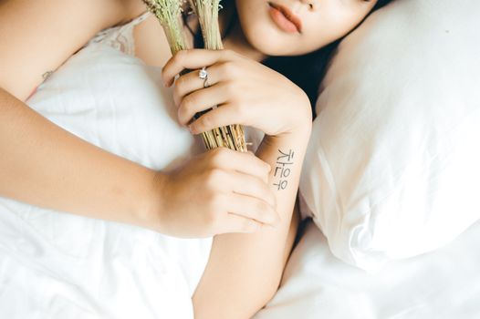High angle of crop female with Chinese hieroglyph tattoo on hand holding bouquet of dried plants while relaxing in comfortable bed in morning