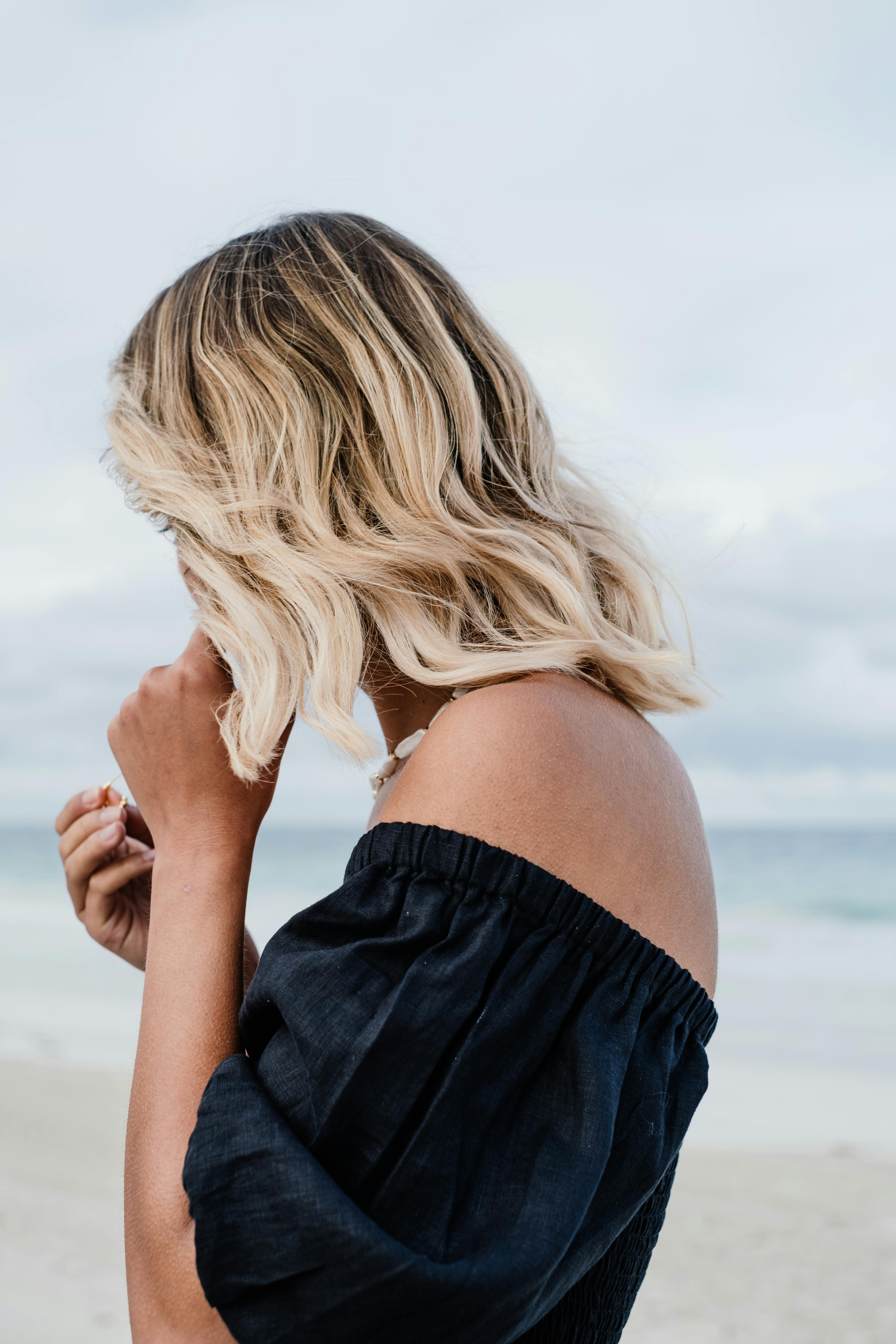 A woman with blonde hair on a beach wearing a black off-shoulder blouse.