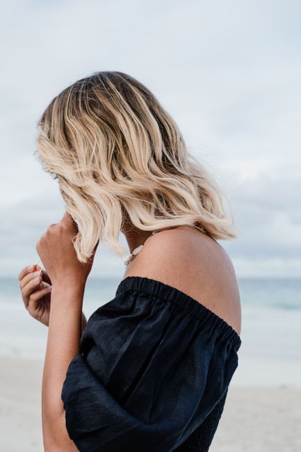 A woman with blonde hair on a beach wearing a black off-shoulder blouse.