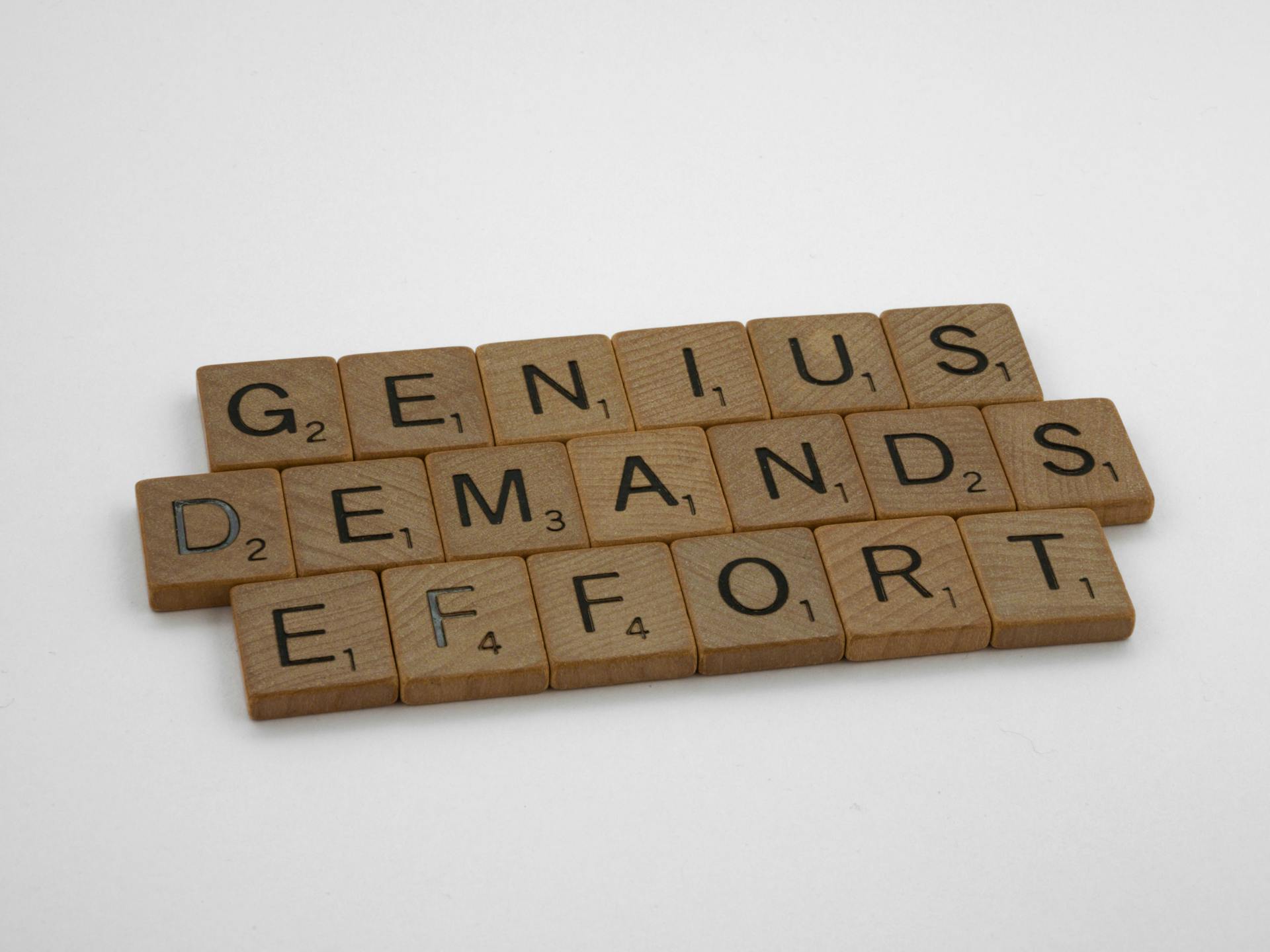 Scrabble tiles spelling 'Genius Demands Effort' on white surface, highlighting creativity and persistence.