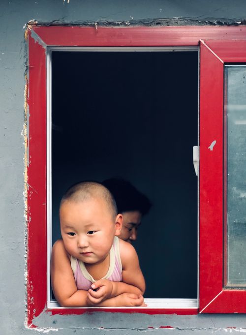 Free A young child with a shaved head gazes out a window with a red frame, displaying curiosity. Stock Photo