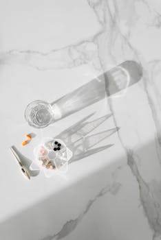 An overhead flat lay of various pills and a glass of water casting shadows on a marble surface.