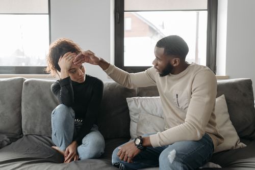 Free Man comforting his unwell partner on a sofa, showing concern and care indoors. Stock Photo