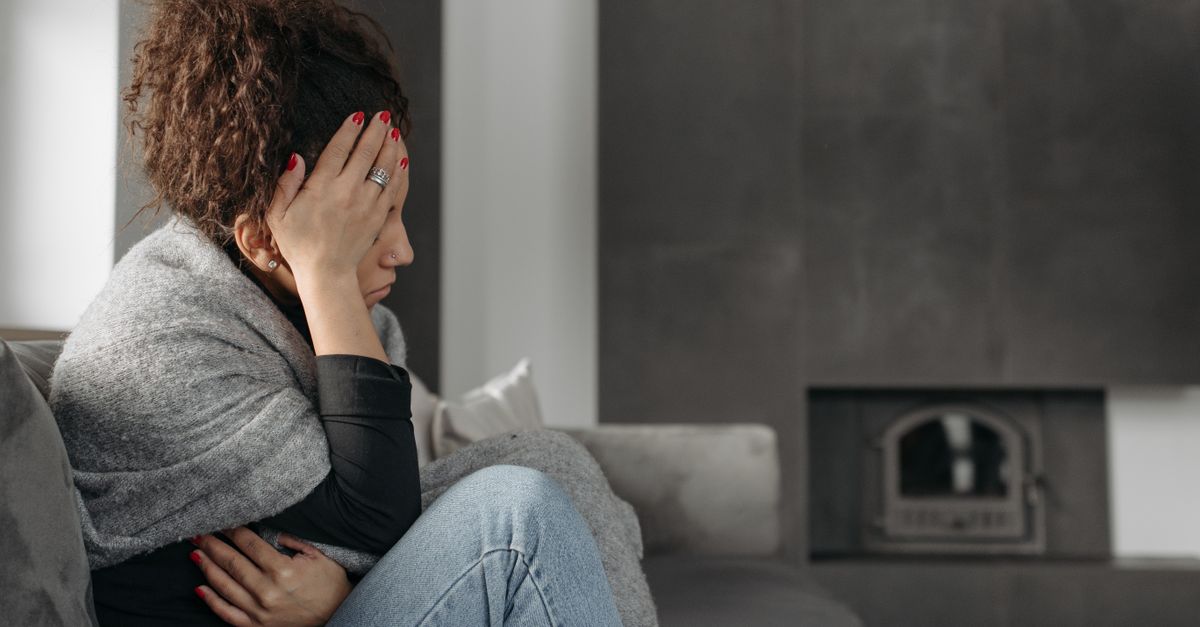 A woman sits on a couch indoors, wrapped in a blanket, holding her head in discomfort.