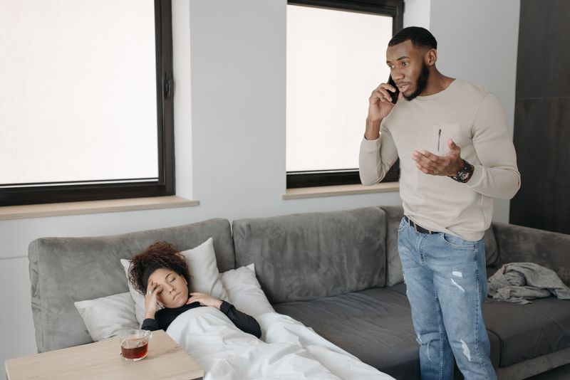A Black man stands in a living room talking on the phone, brow furrowed, while a Black woman lies unwell on the couch under a blanket with a hand to her forehead: the call that decides between urgent care and the emergency room.