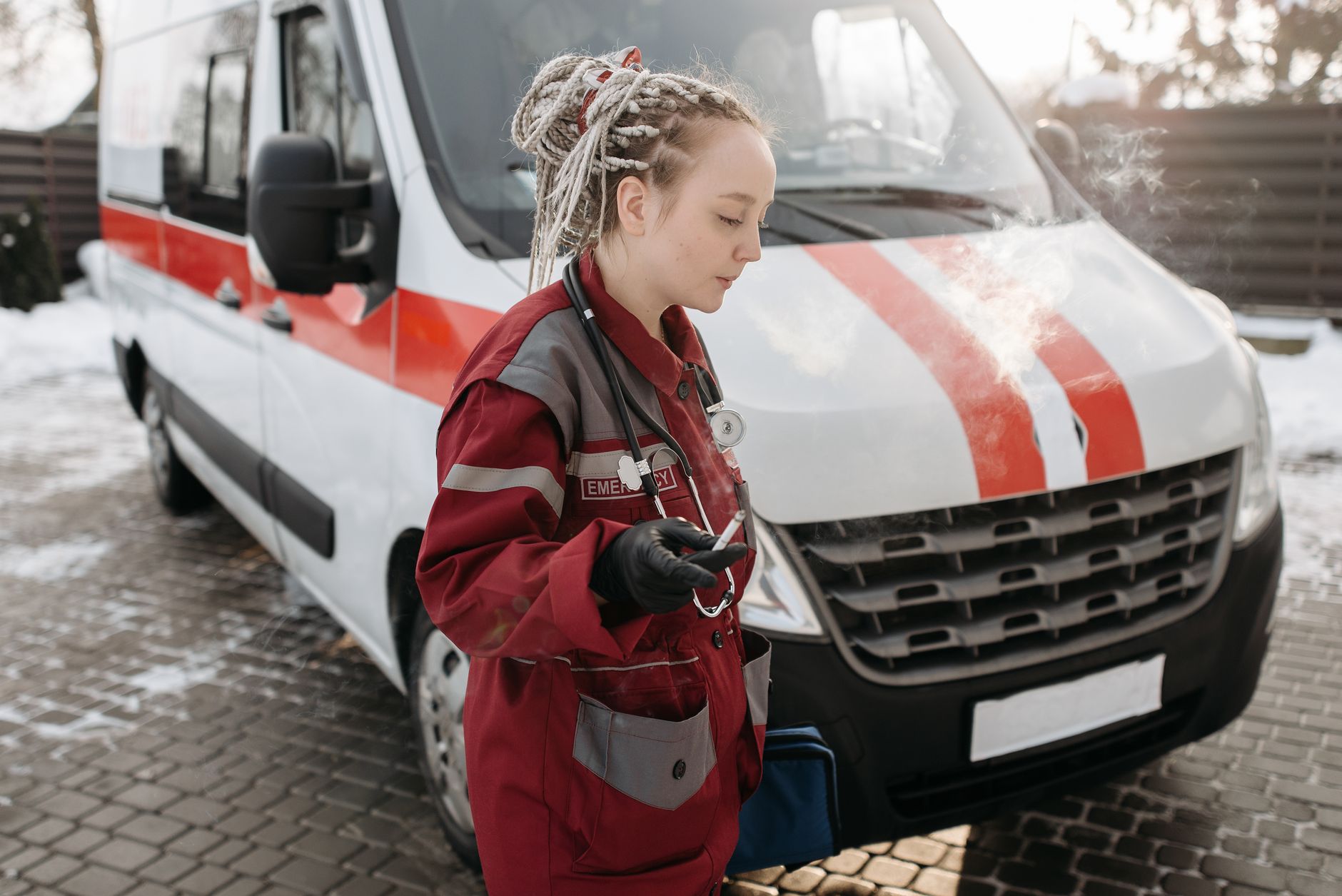 A woman paramedic taking a smoke break beside an ambulance on a snowy winter day.