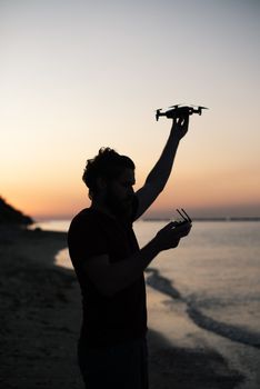Side view of concentrated young bearded male traveler in casual clothes standing on sandy beach and launching drone against picturesque sunset sky
