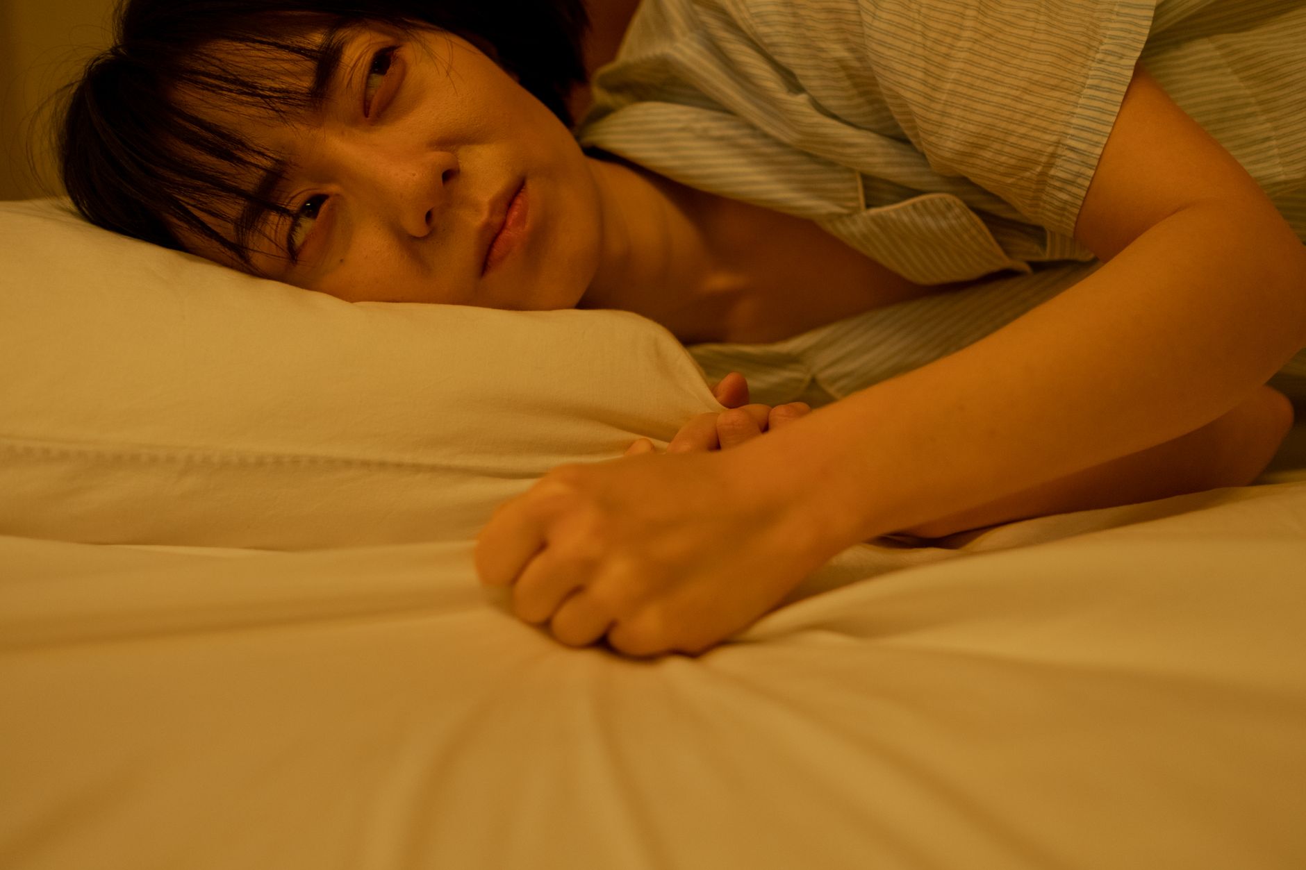 An Asian woman lying on a bed with a thoughtful and melancholic expression, gripping the pillow tightly.