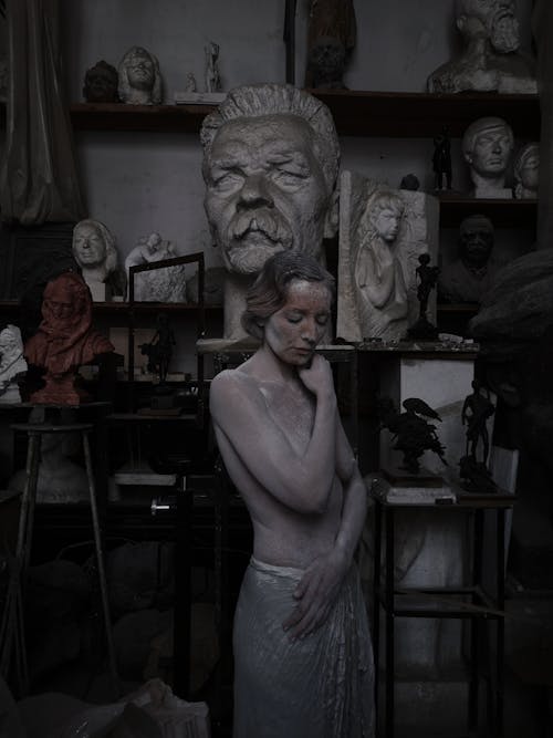 Topless female living sculpture covered with clay looking down while standing near busts and creative clay artworks in modern pottery
