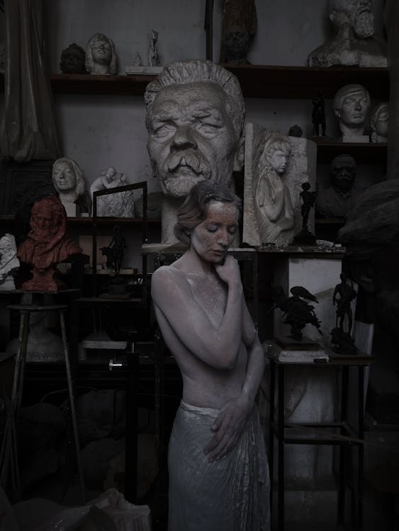 Topless female living sculpture covered with clay looking down while standing near busts and creative clay artworks in modern pottery