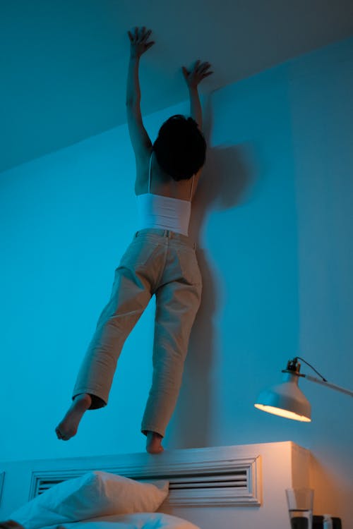 A Woman in White Tank Top Standing on the Bed while Reaching the Ceiling