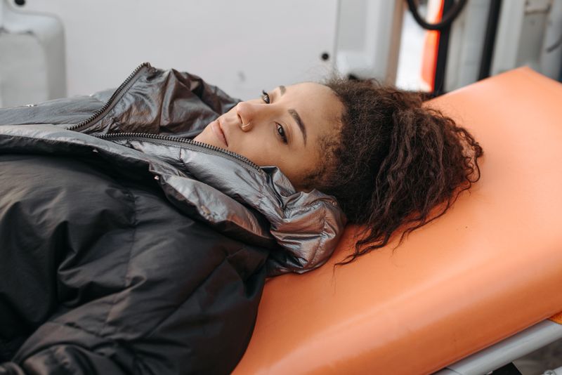 A young Black woman with curly hair lies on her back on an orange ambulance stretcher, wrapped in a dark puffer jacket, looking toward the camera.