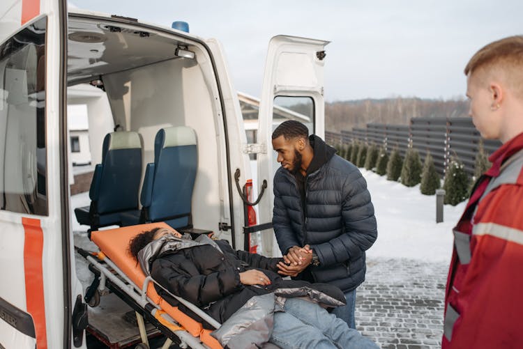 A Man Holding Hands Of His Wife Lying On A Stretcher