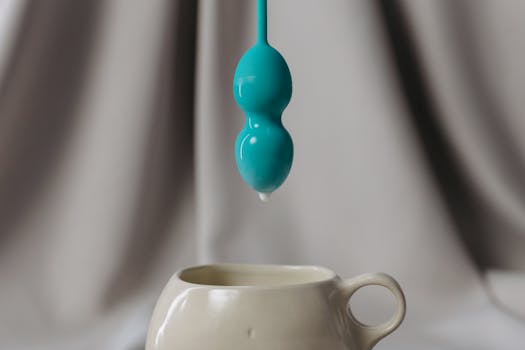 Teal object dripping liquid into an off-white ceramic cup over a blurred background.
