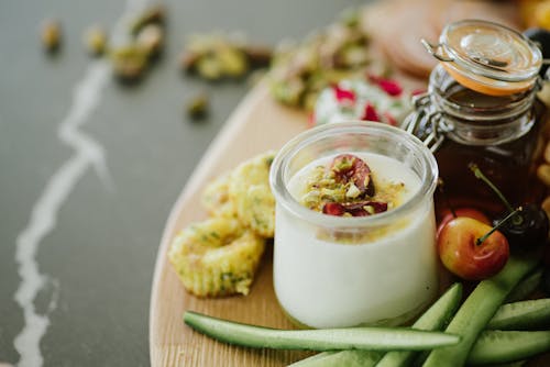 High angle of delicious yogurt with decorative ingredients in glass near cucumber slices and cherries near jar with honey on table in light room