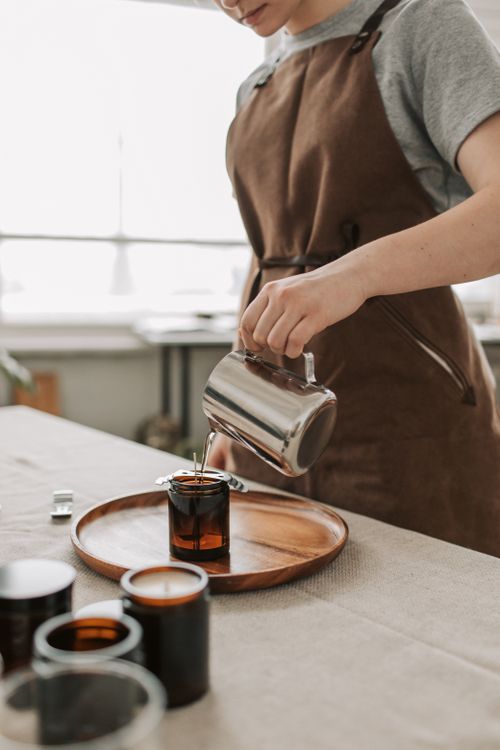 Free Craftsman in apron pours hot wax into a brown glass jar for candle making indoors. Stock Photo