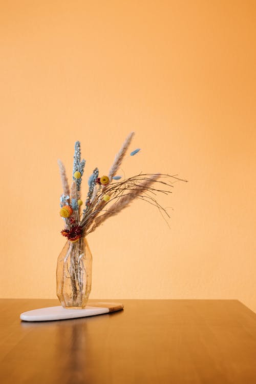 Room with wooden table with glass vase with dried flowers and plants on stand near yellow wall