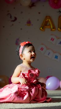 Full body side view of cheerful African American girl in pink dress and headband sitting on floor in light room with garland during holiday celebration