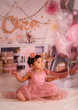Cute baby girl in pink dress celebrates her first birthday with balloons and gifts.