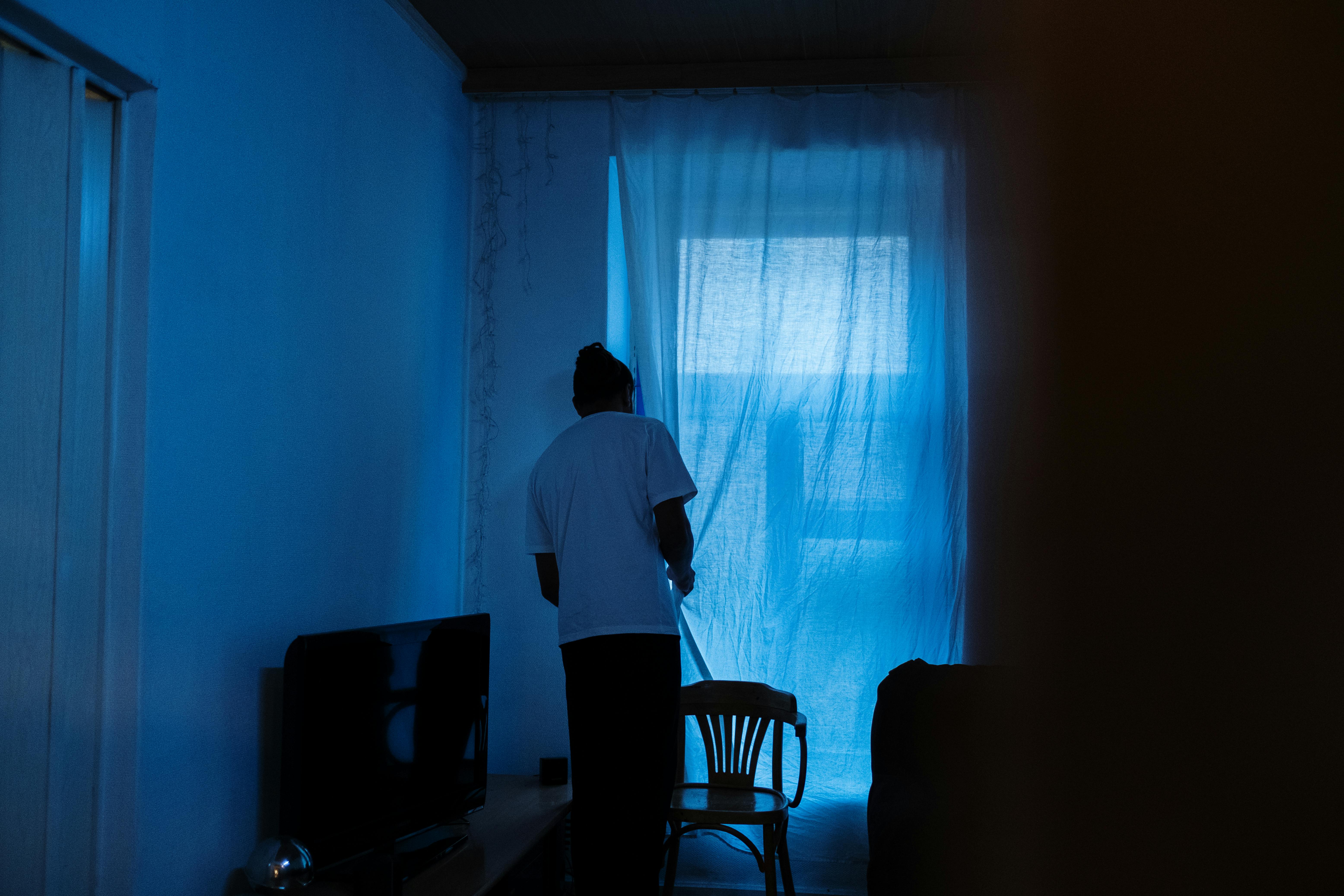 A person stands by a window, creating a silhouette in a dimly lit room. The scene conveys a mood of introspection and solitude.