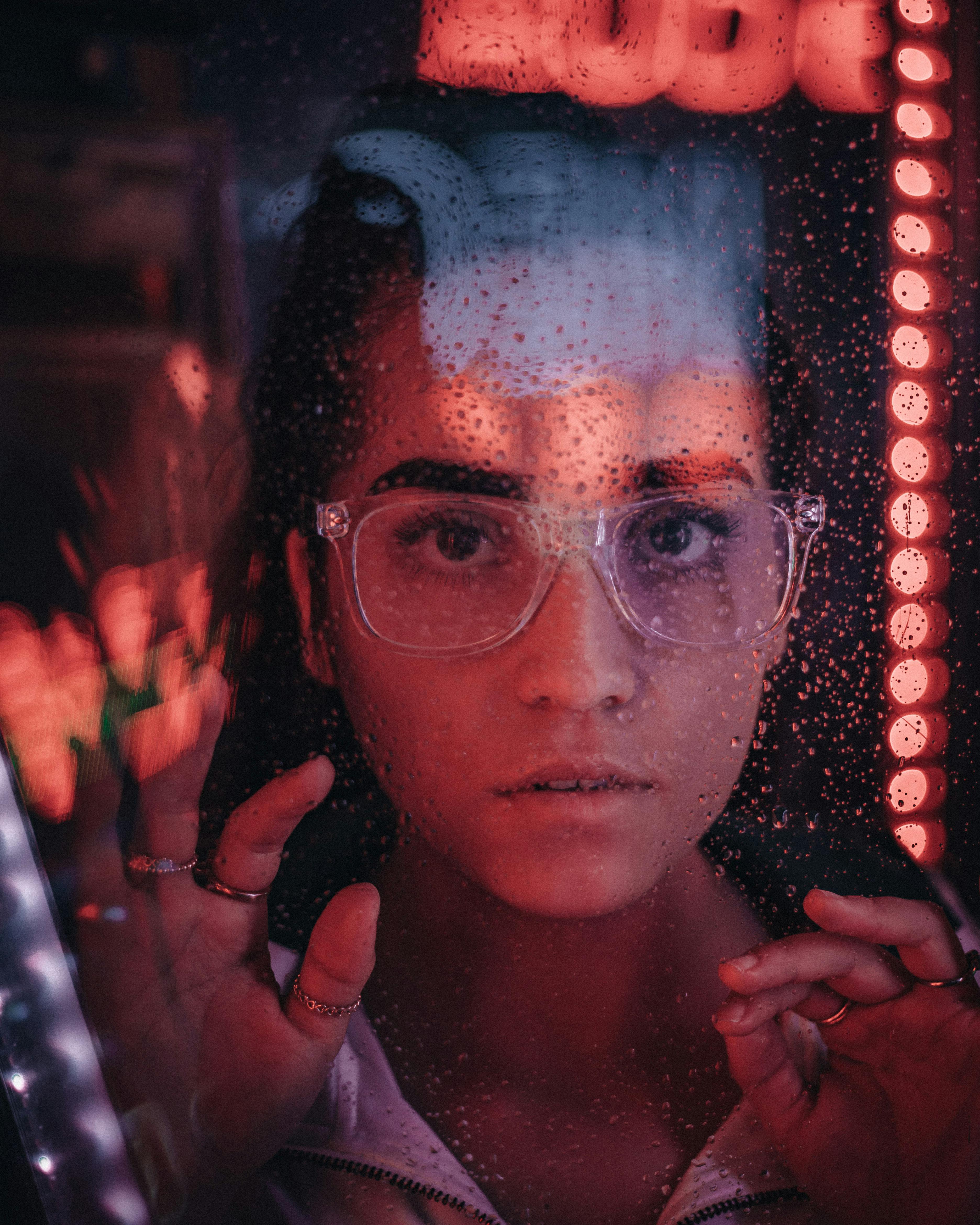 Close-up of a woman with eyeglasses looking through rain-soaked glass in a vibrant setting.