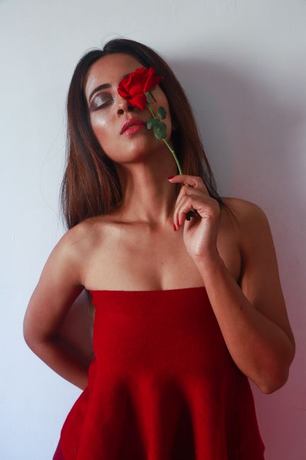 Young woman in red dress with bare shoulders and chest with long straight hair closed eyes and pink lips covering left eye with red rose while standing on white background
