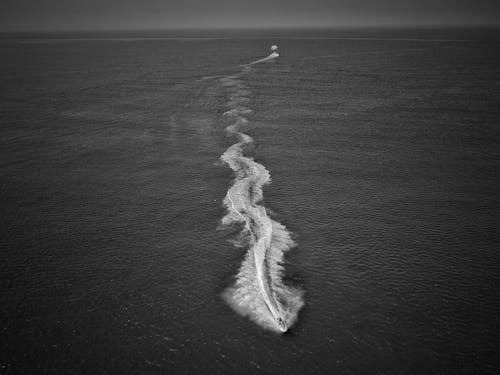 Black and white drone view of fast motorboat sailing in deep dark endless sea and forming waves and splashes in daylight