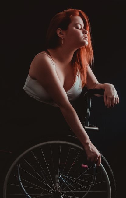 A redhead woman expresses emotion while seated in a wheelchair on a dark backdrop.