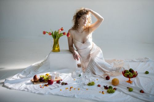 Free Young tender female in white dress touching hair among different sweet fruits on fabric Stock Photo