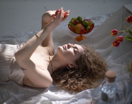 From above of young gentle female lying on white fabric near vase with tasty fruits and blooming red tulips
