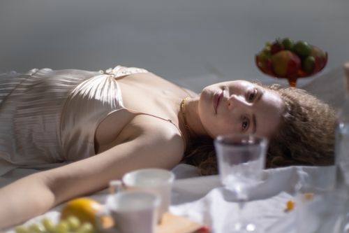 Free Pleasant young female looking at camera and lying on white textile with vase full of fruits composed with glassware Stock Photo