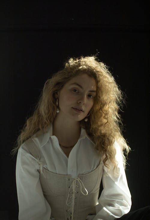 Young female with long curly hair in white shirt and corset looking at camera on black background
